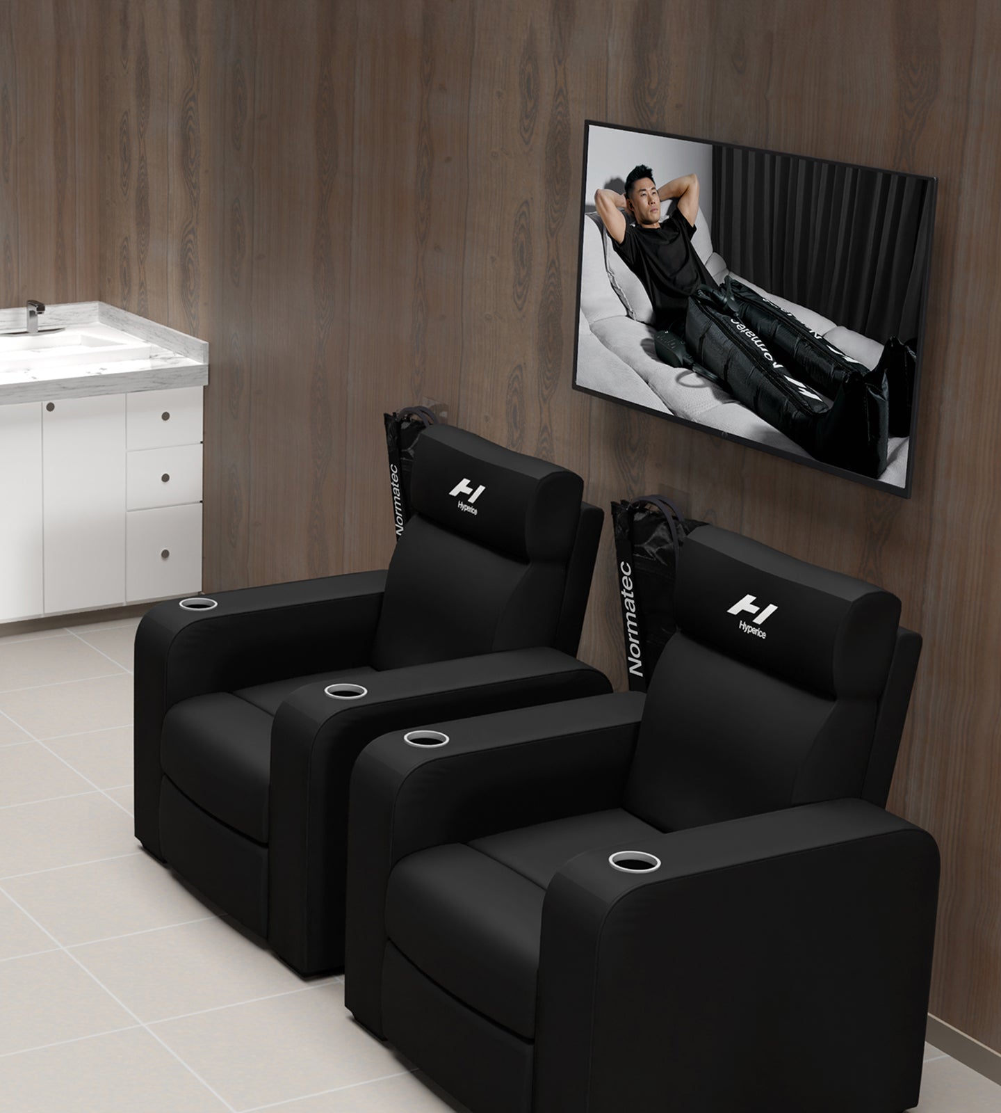 Black Hyperice branded recliner chairs in a room with a TV displaying a person on a couch using their normatec 3 device.