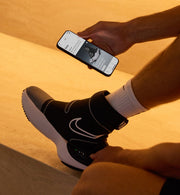 Person wearing Nike x Hyperice Hyperboot on a warm-toned background, holding a smartphone.